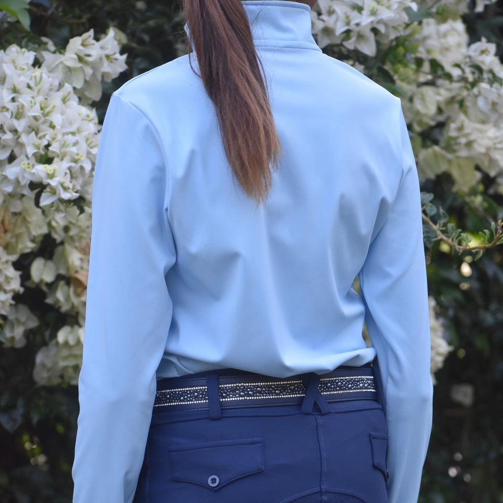 Rear view of a girl wearing navy blue riding tights and a light blue riding base layer with a belt, standing near a bush with white flowers.