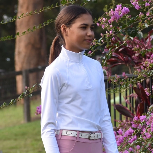 Girl in a white horse riding base layer and pink riding tights standing outdoors with flowers and greenery in the background